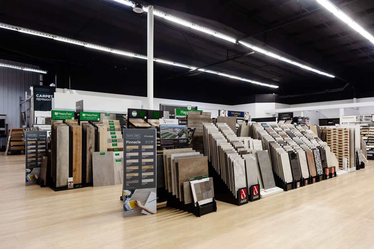 A retail showroom featuring a variety of flooring display racks with tiles, carpets, and laminate samples organized in neat rows. Bright lighting highlights the products, with a clean and spacious layout.